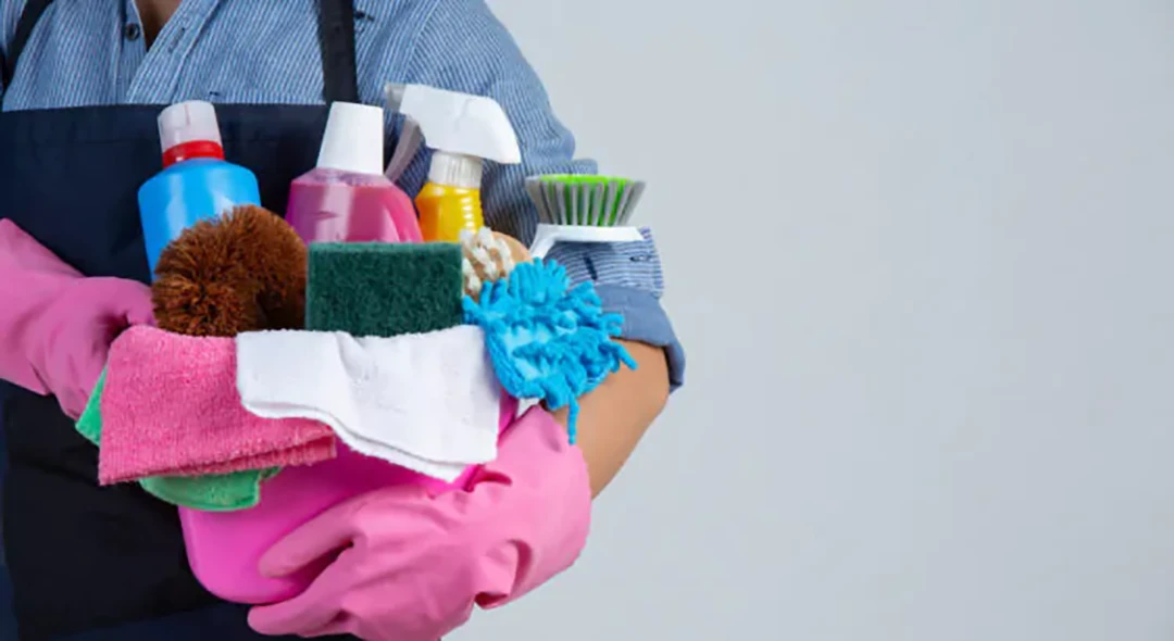Profissional segurando um balde com vários produtos de limpeza doméstica.